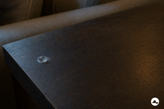 Opgedroogde secondelijmdruppel met witte waas op een donkerhouten bijzettafel