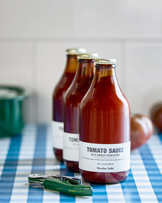 Drie flessen tomatensaus op een blauw-wit geruite doek naast een groene peper.