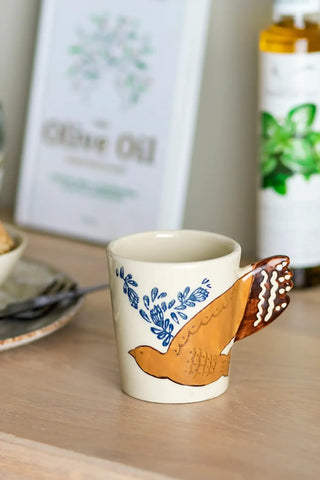 Naturelkleurige Hezha-mok met blauwe takjes en een okerkleurige vogel op een houten aanrecht.