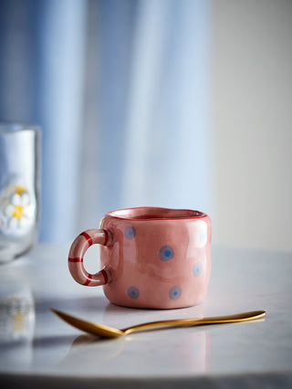 Roze kopje met blauwe stippen en rode rand op een licht tafelblad naast een goudkleurige lepel.
