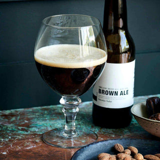 Donkerbruine ale met schuim in een helder bierglas, naast een fles en schaaltjes met noten