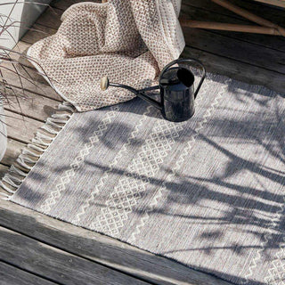 Grijs buitenvloerkleed met wit patroon en franjes op een houten terras, naast een zwarte gieter