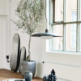 Grijze hanglamp met glazen kap boven houten tafel, naast olijfboom in betonnen pot