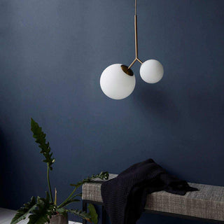 Hanglamp met twee witte bolle kapjes en messingkleurige arm, boven bank met donkere plaid en plant