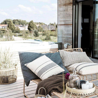 Rieten loungefauteuil met gestreepte en geborduurde kussens op houten terras, met dekentje en glazen vaas