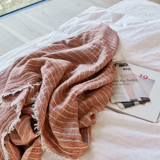 Roestroze gestreepte plaid Alice met franjes, gedrapeerd op een bed naast een tijdschrift