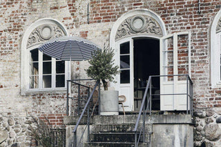 Lichtgrijze Pot Hook bloempot met onderzetter en boom op terras bij een gestreepte parasol