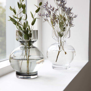 Twee glazen vazen met groene takken en paarse bloemen op een witte vensterbank