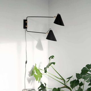 Club Double wandlamp aan een lichte muur boven groene planten, met zichtbaar snoer en schakelaar