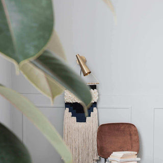 Messingkleurige wandlamp boven een wandkleed, met grote plantbladeren en stoel op de voorgrond