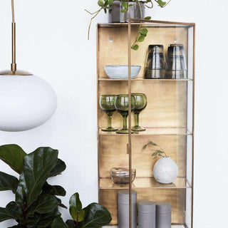Messingkleurige wandvitrine met glazen deur, servies, vazen en glazen aan een witte muur