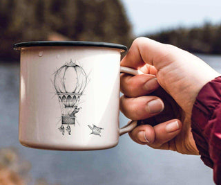 Ligarti emaille mok Ballonvaarders (300 ml) in de hand aan het water, met een fijngetekende luchtballon op de witte mok.
