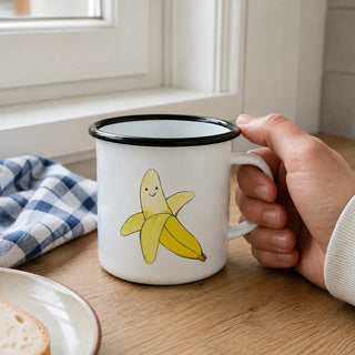 Ligarti emaille mok banaan – 300 ml van dichtbij in gebruik, met de illustratie en dunne zwarte emaille rand zichtbaar.