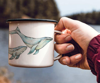 Ligarti emaille mok Blauwe walvis (300 ml) in de hand aan het water, met blauwe walvisillustraties op de witte mok.