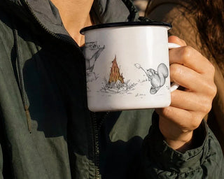 Ligarti emaille mok De Kampvuurclub (300 ml) in de hand, in een uitsnede van de gebruiksfoto, met kampvuurillustratie zichtbaar.