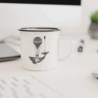 Witte emaille mok met zwarte rand en walvis met luchtballon op een bureau