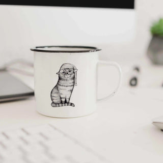 Witte emaille mok met kattenillustratie en slaapmuts op een bureau naast een laptop
