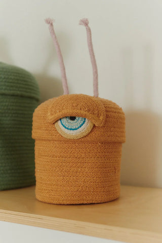 Oranje alienmand met één blauw oog en roze antennes op een plank.