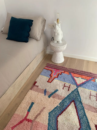 1001 Nights-vloerkleed met roze en blauwe geometrische motieven naast een bed.