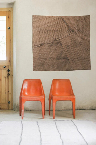 Bruin Stoashed-wandtextiel boven twee oranje stoelen en een lichtgrijs Stoashed-vloerkleed op de vloer.