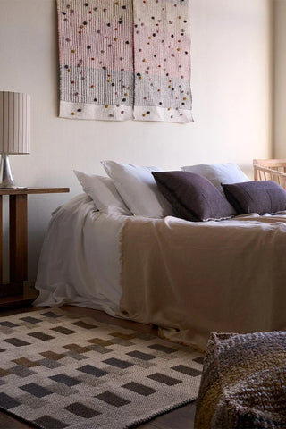 Naturelkleurige Intersecting-vloerkleed met blokmotieven naast een bed.