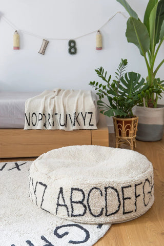 Naturel ABC-poef met zwarte letters op een ABC-vloerkleed bij een bed.