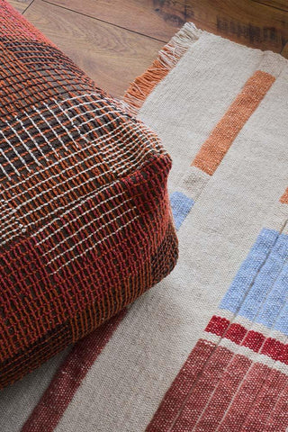 City Maple Red-poef met geweven zijde op een vloerkleed met gekleurde banen.