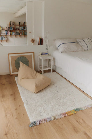 Licht honingkleurige Cleo-zitzak op een vloerkleed naast een bed.