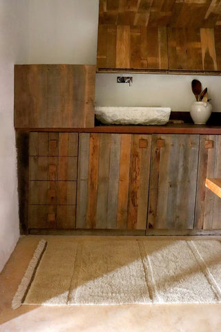 Beige Steppe-vloerkleed met lichte franjeranden bij een houten badkamermeubel.