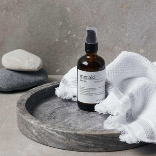 Meraki body oil in bruine fles met spraydop op stenen dienblad met witte handdoek en stenen