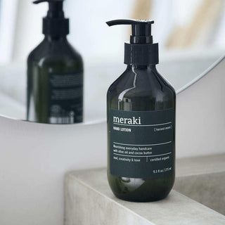 Meraki handlotion Harvest Moon in donkergroene pompflacon op een licht stenen blad voor een spiegel