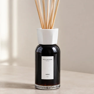 Detail van Millefiori Milano Reed Diffuser 250 ml Nero met donker glas, witte kraag en natuurlijke geurstokjes in raamlicht.