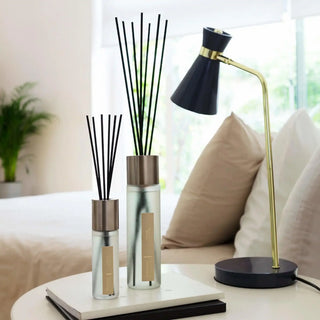 Twee glazen geurdiffusers met zwarte stokjes en messing doppen op nachtkastje naast blauwe tafellamp