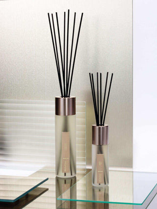Twee glazen geurdiffusers met zwarte stokjes en roze metalen doppen op glazen tafel