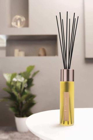 Gele geurdiffuser met zwarte stokjes en metalen dop, geplaatst op witte tafel in moderne woonkamer