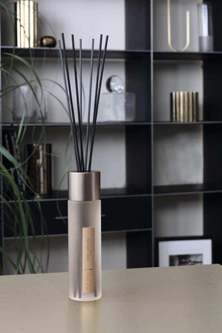 Beige geurdiffuser met zwarte stokjes en metalen dop, geplaatst op tafel in moderne woonkamer