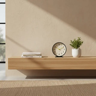 Newgate Q tafelklok zwart met ronde lichte wijzerplaat en goudkleurige wijzers, op een zwevende wandplank naast boeken en een plant
