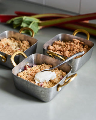 Drie metalen mini braadschalen met goudkleurige handgrepen, gevuld met een kruimelig gerecht