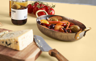 Ovale mini pan met olijven, tomaten en paprika naast kaas op een houten plank