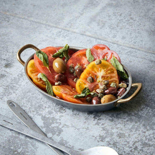 Ovale mini pan gevuld met tomaten, olijven, kappertjes en munt naast bestek