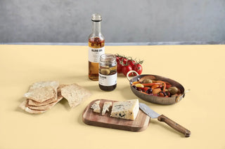 Houten snijplank met blauwe kaas en mes, omringd door crackers, olijven en tomaten