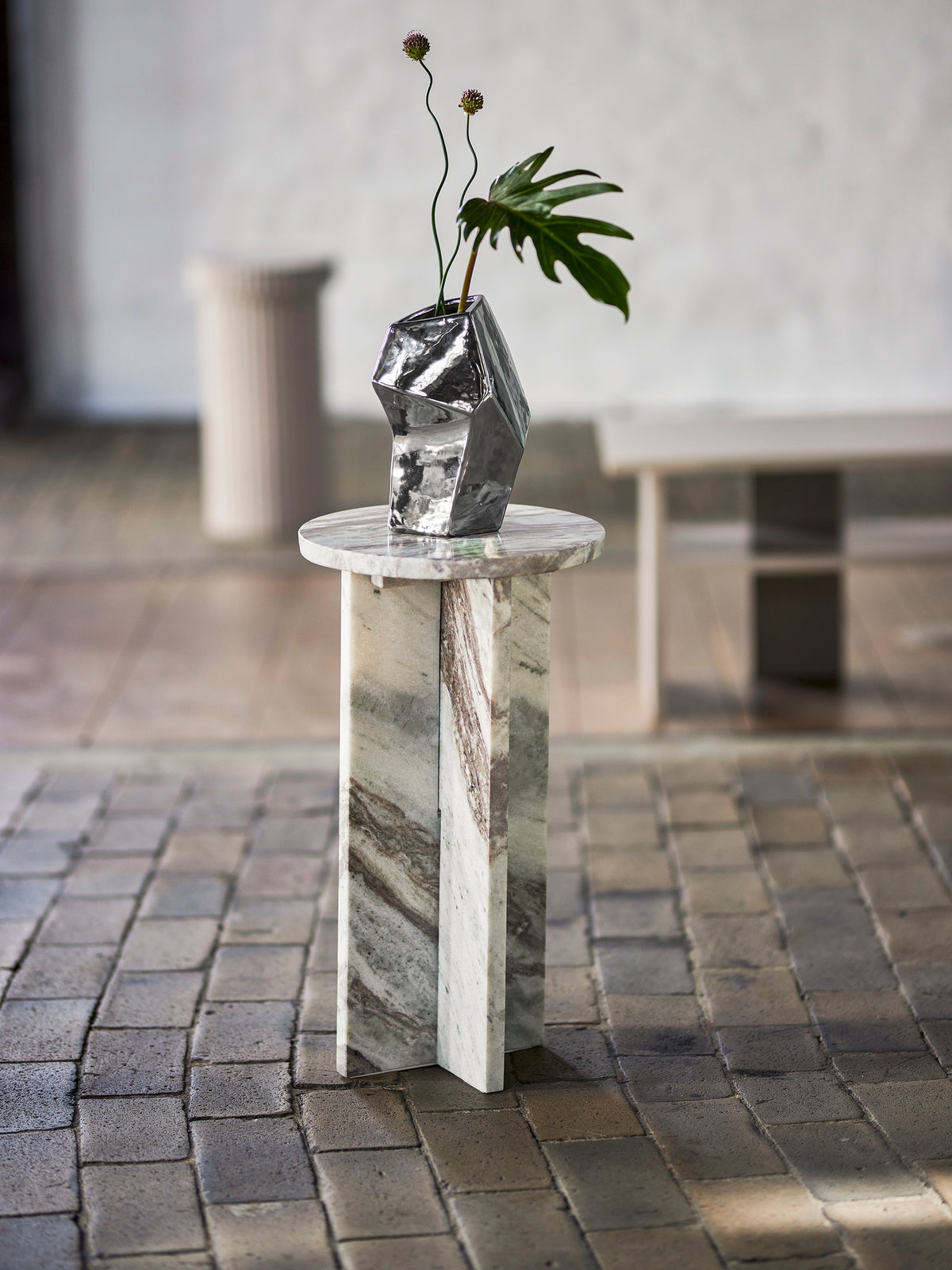 Sølvfarvet geometrisk vase med grønne grene på en høj marmorsokkel i et lyst og roligt rum